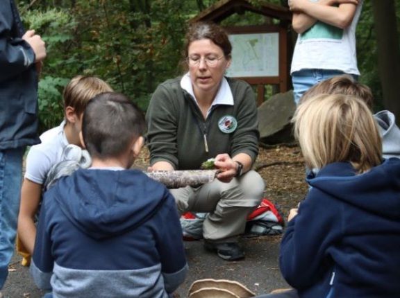 Christine Speck hockt im Wald vor einer Kindergruppe und zeigt ihnen ein Stück Holz mit Pilzen