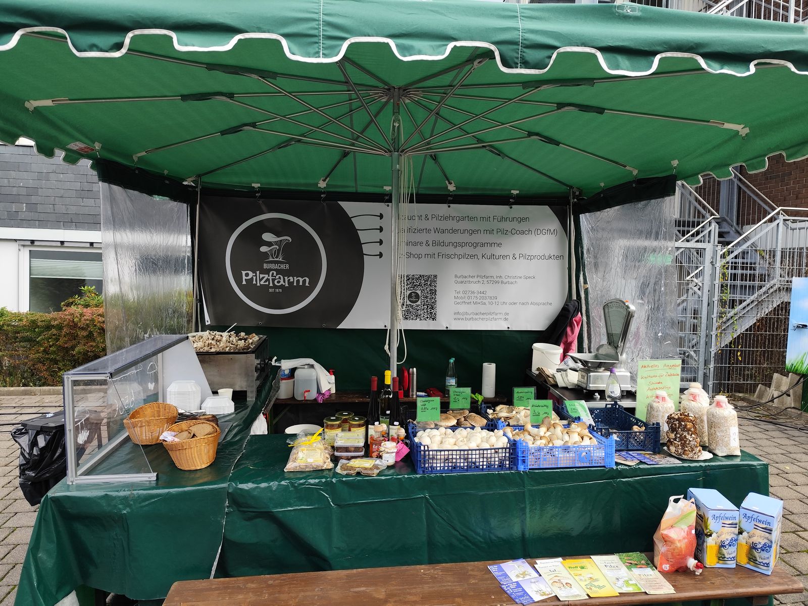 Marktstand der Burbacher Pilzfarm unter einem grünen Schirm, mit Kisten voller Pilze, Gläsern, Zuchtkulturen und dem Banner der Pilzfarm